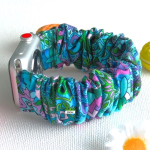 May include: A teal, purple, and green patterned scrunchie watch band. The band is made of fabric and is attached to a silver Apple Watch. The word "Shirley" is printed on the band.