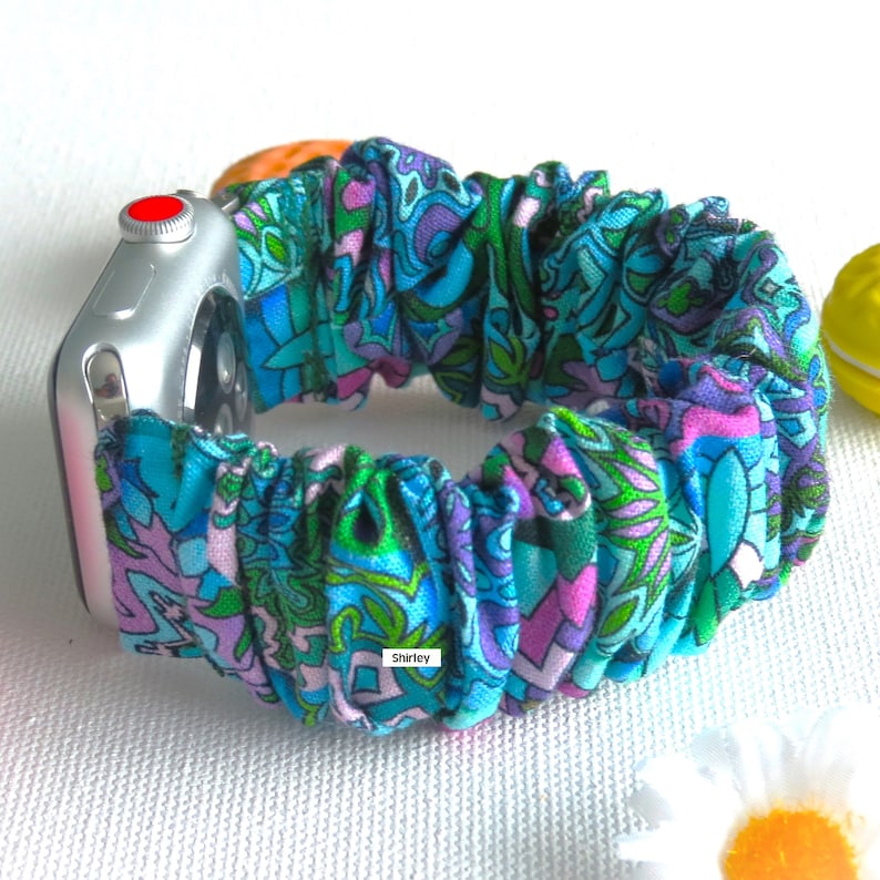 May include: A teal, purple, and green patterned scrunchie watch band. The band is made of fabric and is attached to a silver Apple Watch. The word "Shirley" is printed on the band.