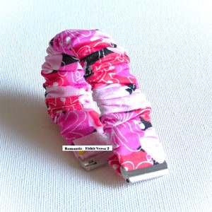 May include: A pink, black, and white patterned fabric watch band for a Fitbit Versa 2. The band is made of a soft, stretchy material and has a secure clasp.
