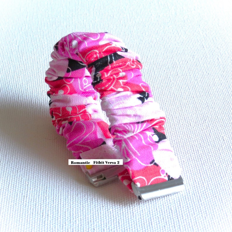 May include: A pink, black, and white patterned fabric watch band for a Fitbit Versa 2. The band is made of a soft, stretchy material and has a secure clasp.
