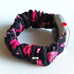 May include: Black and pink fabric watch band with a heart pattern. The band is elastic and fits around a silver Apple Watch. The band has the text "XOXO" printed on it.