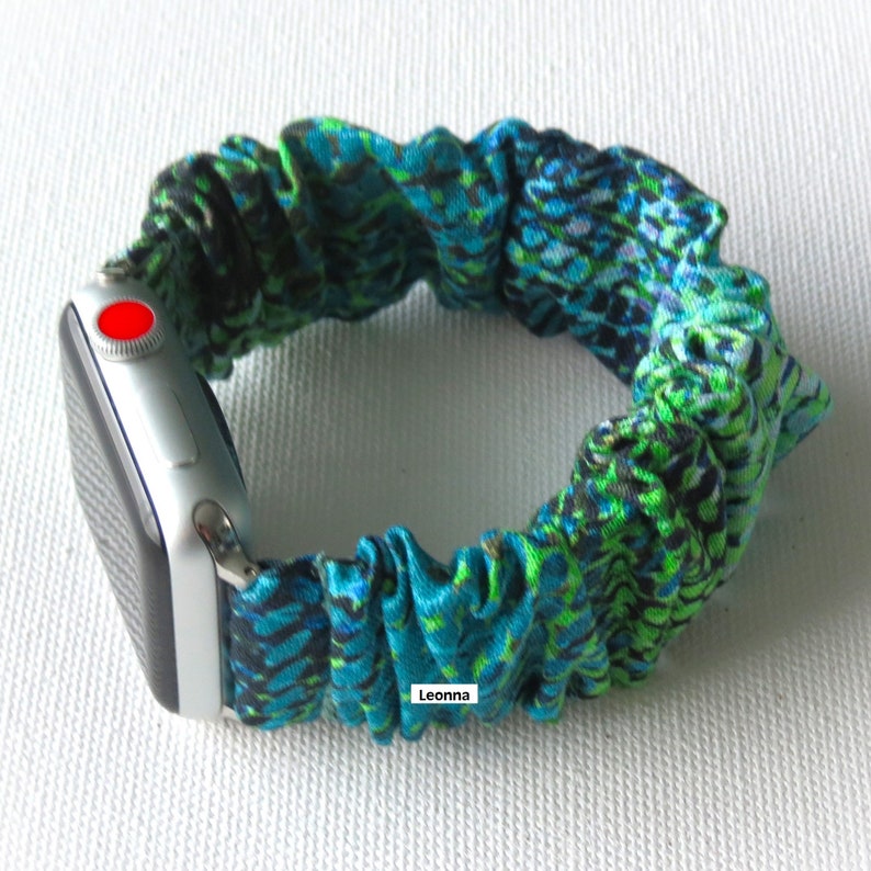 May include: A teal and green patterned scrunchie watch band attached to a silver Apple Watch. The scrunchie band has a repeating design of teal and green shapes. The watch has a red button on the side. The word "Leonna" is printed on the band.