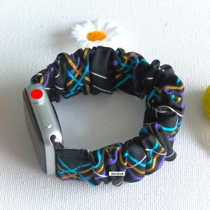 May include: A black scrunchie watch band with a colorful geometric pattern in teal, gold, and purple. The band is attached to a silver smartwatch with a red button. A white daisy is in the background.
