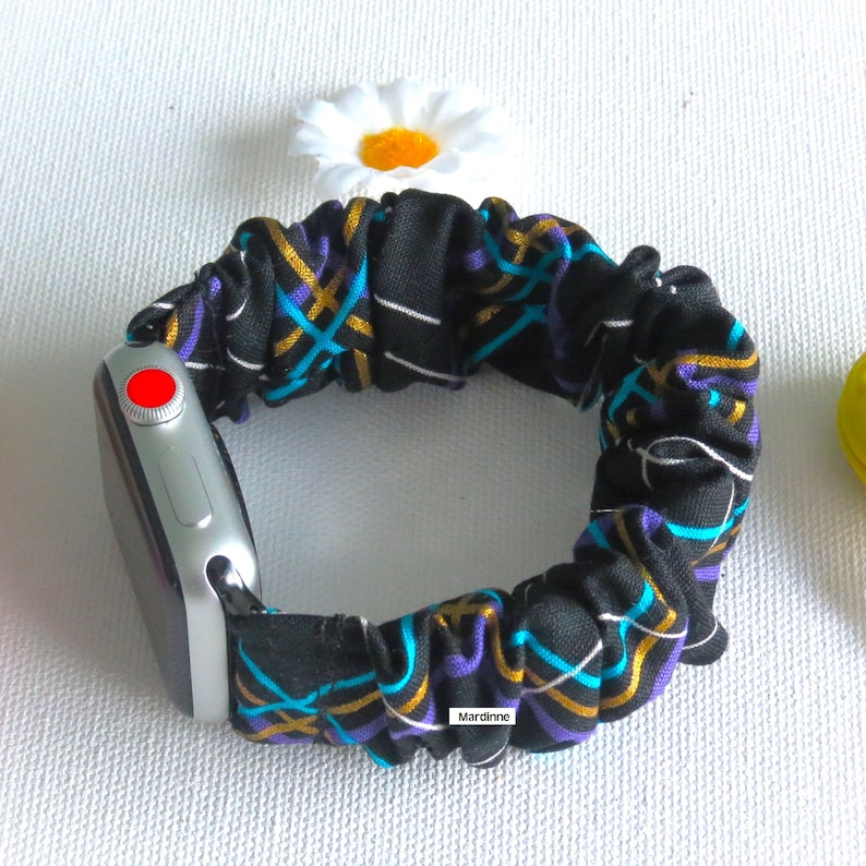 May include: A black scrunchie watch band with a colorful geometric pattern in teal, gold, and purple. The band is attached to a silver smartwatch with a red button. A white daisy is in the background.