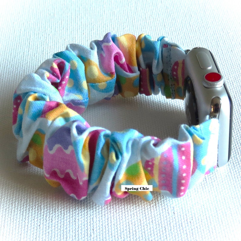 May include: A colorful, patterned fabric Apple Watch band with a blue, pink, yellow, and green design. The band is made of a stretchy, elastic material and has a "Spring Chic" label.