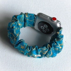 May include: A teal and gold patterned scrunchie watch band attached to a silver Apple Watch. The scrunchie band is made of gathered fabric with a swirling gold design. The watch has a red button and a black screen.