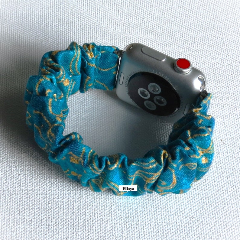 May include: A teal and gold patterned scrunchie watch band attached to a silver Apple Watch. The scrunchie band is made of gathered fabric with a swirling gold design. The watch has a red button and a black screen.