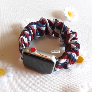 Op de afbeelding: Een rood, wit en blauw gestreept stoffen horlogebandje met een zilveren Apple Watch. De wijzerplaat van het horloge is zwart en het bandje is gemaakt van rekbaar stof.