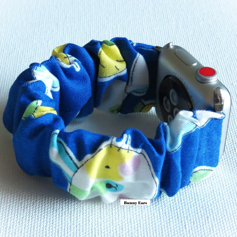 May include: A blue and white patterned watch band with a white bunny design. The band is made of fabric and has a stretchy, elastic feel. The band is attached to a silver Apple Watch.