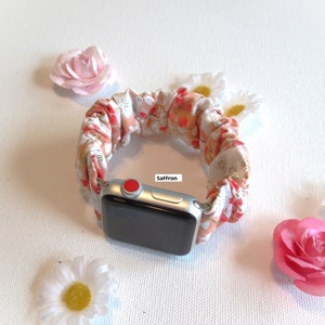 K&ouml;nnte beinhalten: Eine wei&szlig;e Apple Watch mit einem Armband aus Stoff mit Blumenmuster. Das Armband ist im Scrunchie-Stil und hat einen roten Knopf auf dem Zifferblatt der Uhr. Der Text "Saffron" ist auf dem Zifferblatt der Uhr sichtbar.