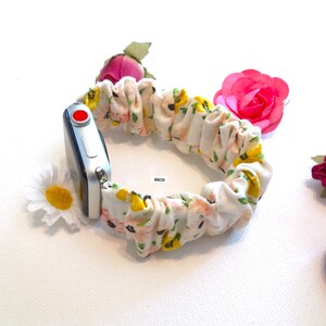 May include: A white Apple Watch with a floral print fabric band. The band is a scrunchie style and has yellow and pink flowers on a white background.