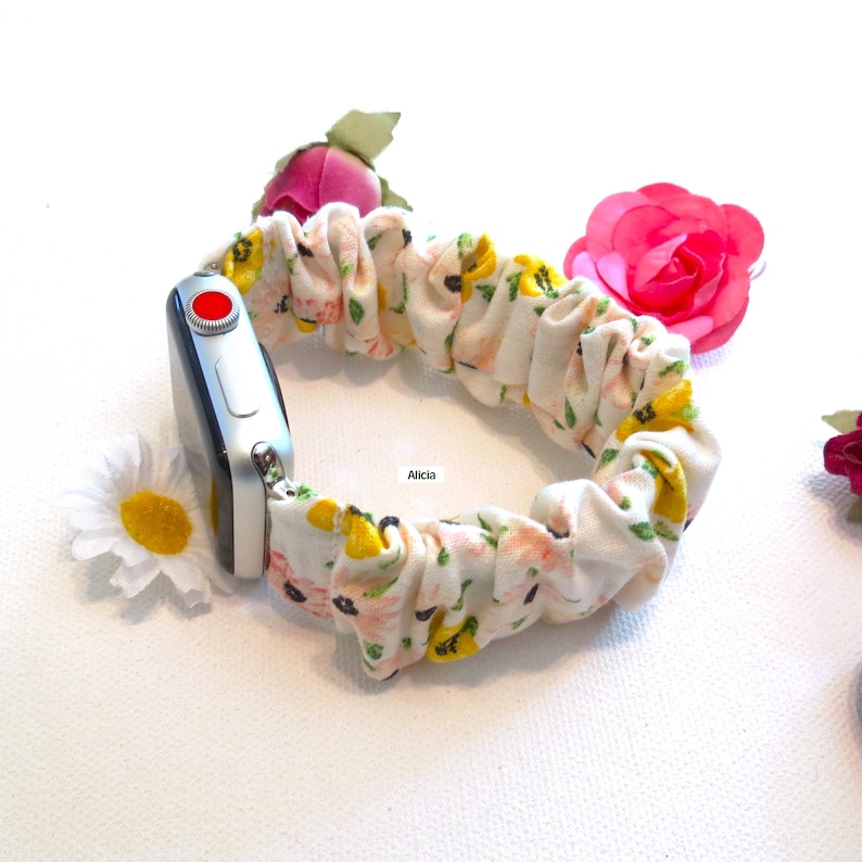May include: A white Apple Watch with a floral print fabric band. The band is a scrunchie style and has yellow and pink flowers on a white background.