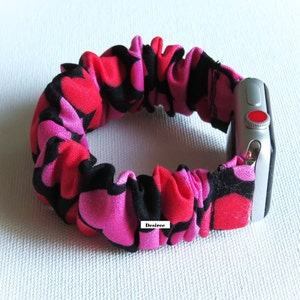 May include: A pink and red heart print watch band with a black background. The band is made of stretchy fabric and is designed to fit a smartwatch.