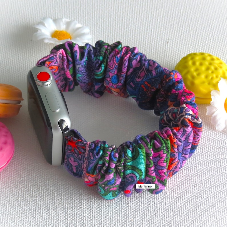 May include: A colorful scrunchie watch band with a paisley pattern in shades of purple, blue, green, and orange. The band is attached to a silver smartwatch with a red button. The word "Marianne" is printed on the band.