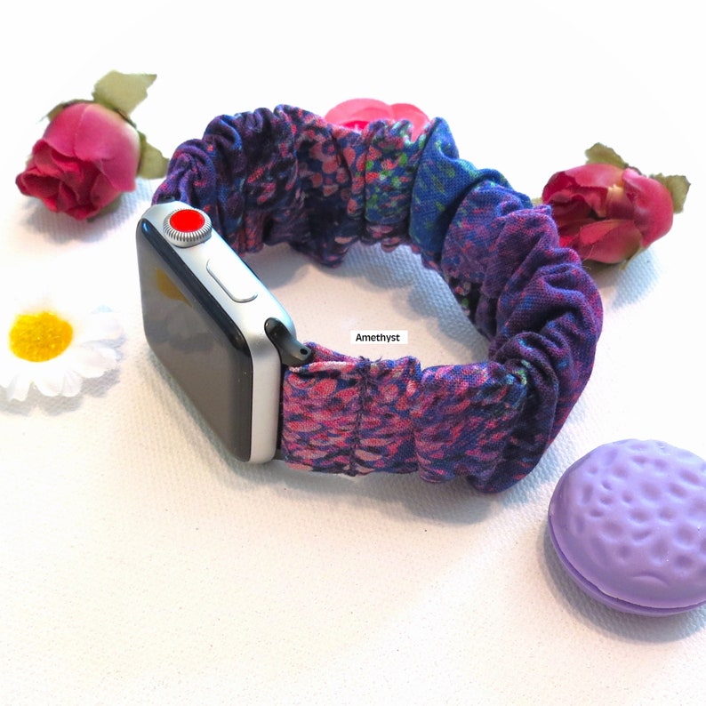 May include: A silver Apple Watch with a patterned scrunchie band in shades of purple, pink, and blue. The band features a floral design and the word "Amethyst". The watch is surrounded by flowers and a lavender-colored object.