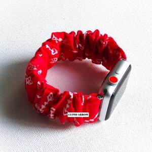 May include: Red and white patterned fabric watch band with a silver Apple Watch. The band is elastic and has the words "CUPID ARROW" printed on it.
