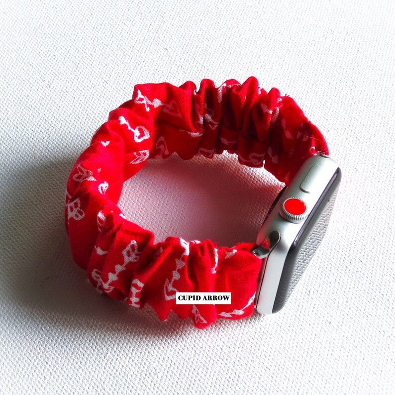 May include: Red and white patterned fabric watch band with a silver Apple Watch. The band is elastic and has the words "CUPID ARROW" printed on it.