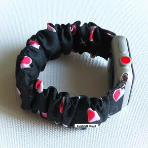 May include: Black and pink heart patterned fabric watch band for an Apple Watch. The band is made of a stretchy, soft fabric and has a red button on the side of the watch.