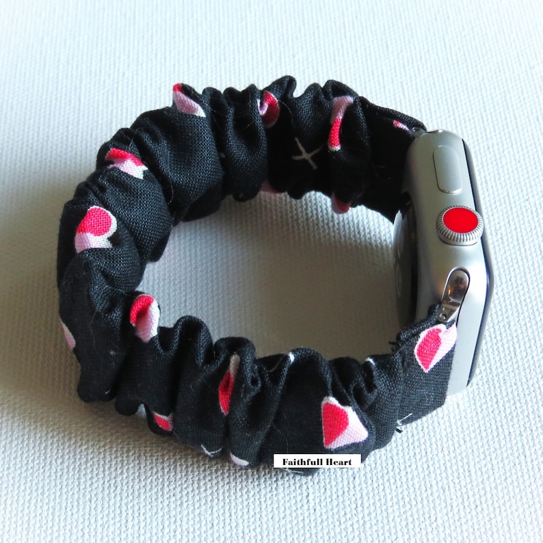May include: Black and pink heart patterned fabric watch band for an Apple Watch. The band is made of a stretchy, soft fabric and has a red button on the side of the watch.