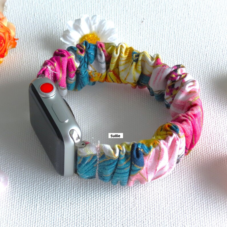 K&ouml;nnte beinhalten: Ein farbenfrohes Armband aus Stoff mit floralem Muster f&uuml;r eine Apple Watch. Das Armband besteht aus einem weichen, dehnbaren Material und hat ein leuchtend pinkfarbenes, gelbes, blaues und wei&szlig;es Design. Das Armband ist auf einer silbernen Apple Watch mit schwarzem Zifferblatt zu sehen.