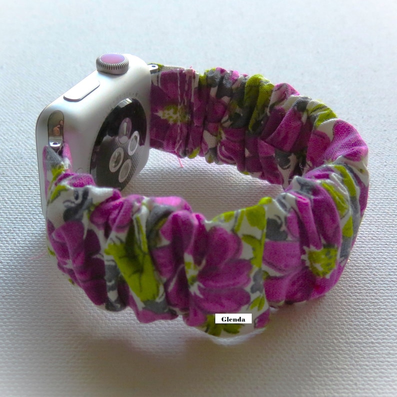 May include: A white Apple Watch with a purple and green floral fabric band. The band is elastic and has a ruffled look. The watch face is black and has a digital display. The text "Glenda" is visible on the band.