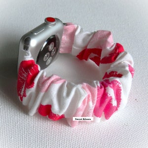 May include: A pink and white Apple Watch band with a red lip print. The band is made of a soft, stretchy fabric and is designed to fit snugly around the wrist. The band is shown on a silver Apple Watch.