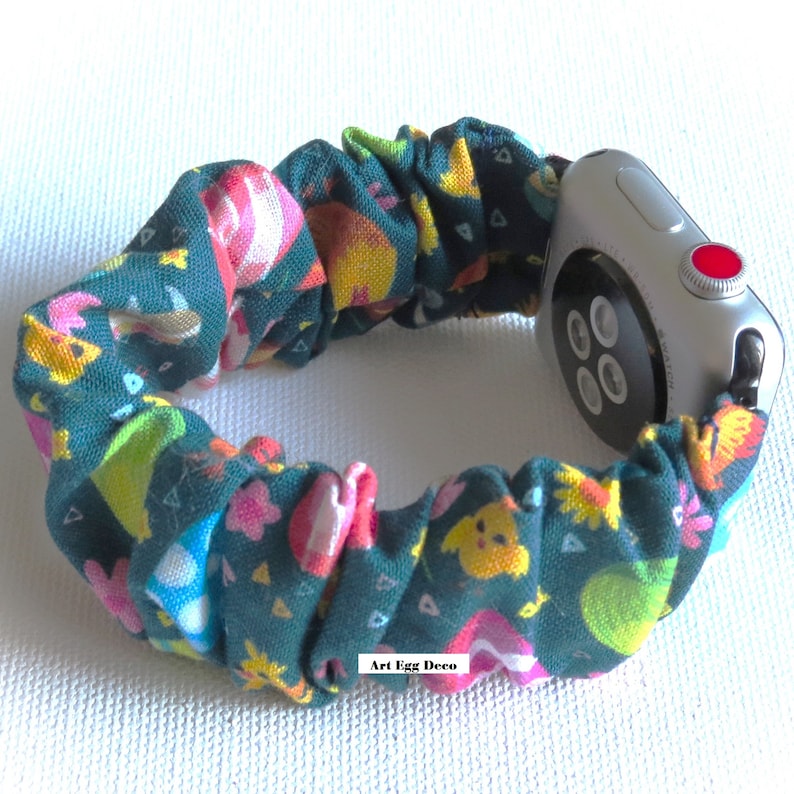 May include: A green, white, pink, and yellow patterned fabric Apple Watch band with a silver Apple Watch. The band is made of a stretchy, elastic material and has a fun, whimsical design. The text "Art Egg Deco" is printed on the band.
