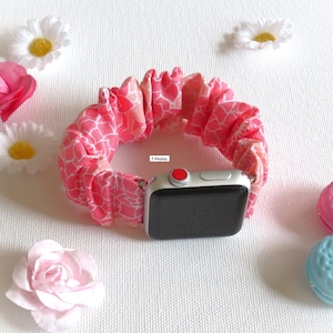 May include: A pink and white patterned fabric watch band for an Apple Watch. The band is made of a soft, stretchy material and has a ruffled look. The text "7 Peona" is visible on the band.