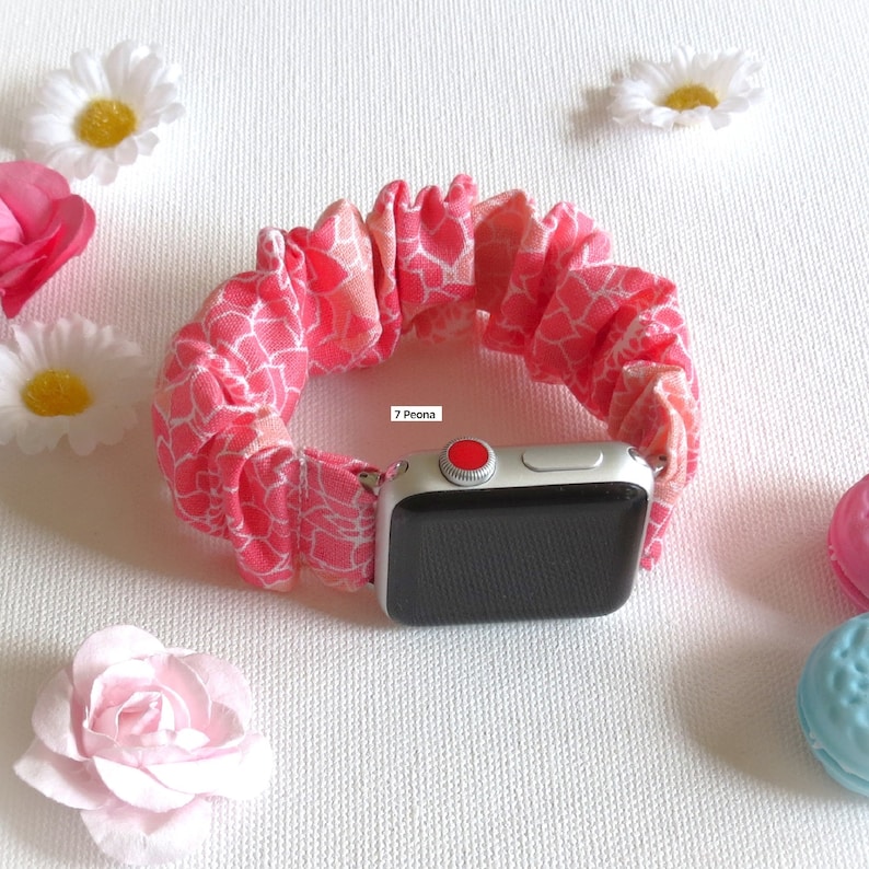 May include: A pink and white patterned fabric watch band for an Apple Watch. The band is made of a soft, stretchy material and has a ruffled look. The text "7 Peona" is visible on the band.