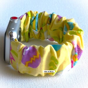 May include: A yellow Apple Watch band with an Easter egg pattern. The band is made of a stretchy fabric and has a red button on the side. The text "Chic & Bows" is visible on the band.