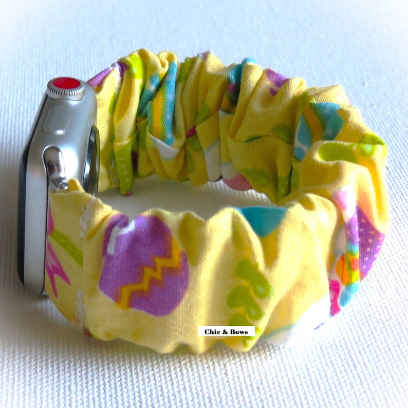 May include: A yellow Apple Watch band with an Easter egg pattern. The band is made of a stretchy fabric and has a red button on the side. The text "Chic & Bows" is visible on the band.
