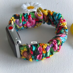 May include: A colorful floral patterned fabric watch band for an Apple Watch. The band is made of a stretchy, elastic material and has a red button on the side. The band is attached to a silver Apple Watch.