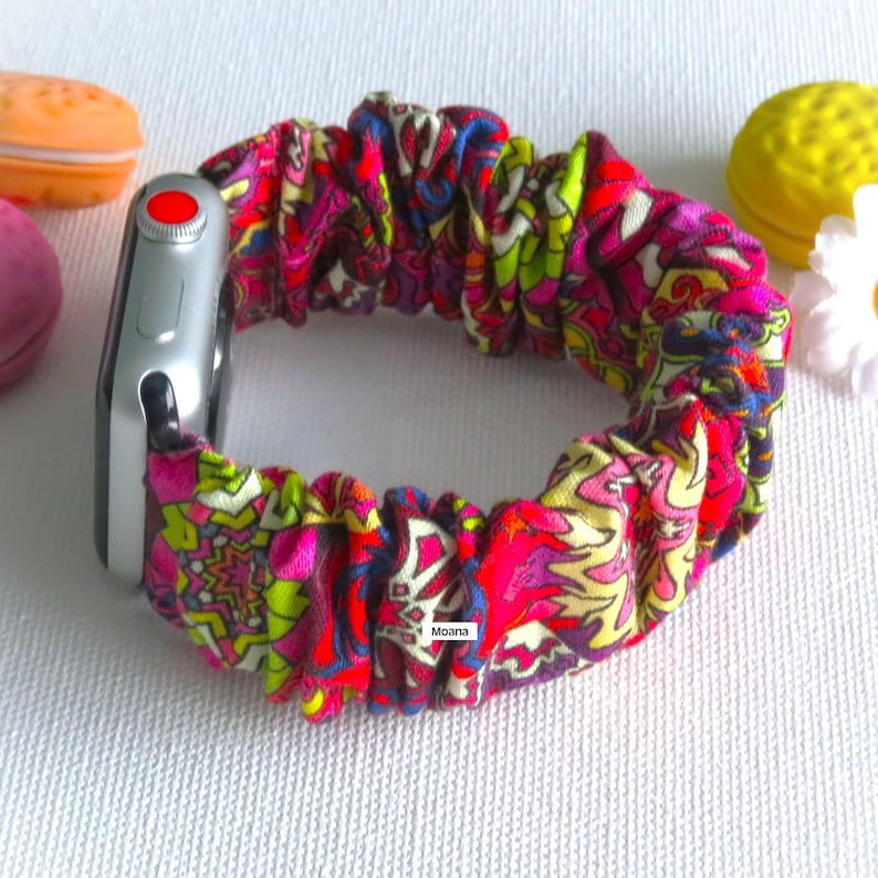 May include: A colorful scrunchie watch band with a floral pattern in shades of pink, red, yellow, and green. The band is attached to a silver Apple Watch. The word "Moana" is visible on the band.