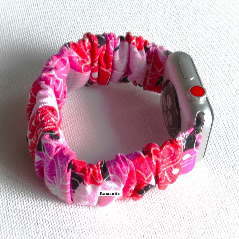 May include: A pink, red, and black floral patterned fabric Apple Watch band. The band is made of a stretchy material and is designed to fit snugly around the wrist. The band is shown on a silver Apple Watch.