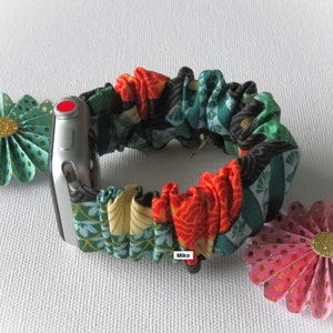 K&ouml;nnte beinhalten: Ein farbenfrohes Armband aus Stoff mit einem gr&uuml;n-orange-schwarzen Muster. Das Armband besteht aus einem weichen, dehnbaren Material und ist f&uuml;r eine Smartwatch konzipiert. Das Armband ist auf einer silbernen Smartwatch mit einem roten Knopf zu sehen.