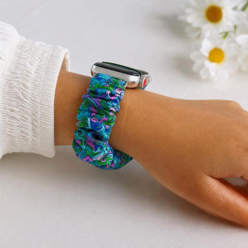 May include: A silver smartwatch with a teal, purple, and green floral patterned scrunchie band. The band is made of a soft, gathered fabric. The watch is on a wrist with a white long-sleeved shirt.