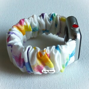 May include: A white Apple Watch band with a tie-dye pattern in shades of pink, yellow, blue, and purple. The band is made of a soft, stretchy fabric and has a "Tie Dye Bunny" label.