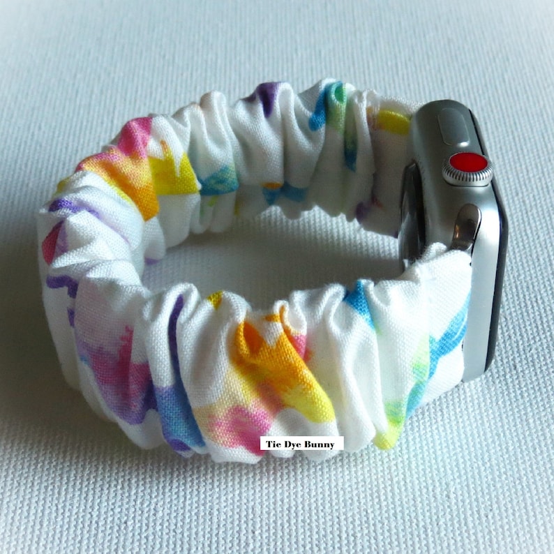 May include: A white Apple Watch band with a tie-dye pattern in shades of pink, yellow, blue, and purple. The band is made of a soft, stretchy fabric and has a "Tie Dye Bunny" label.