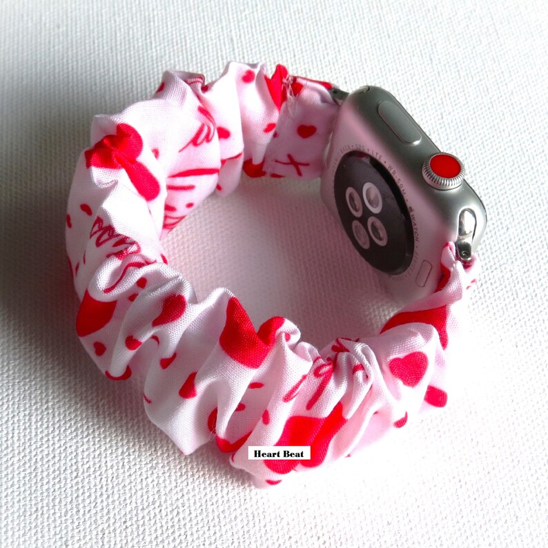 May include: A white and red patterned Apple Watch band with a heart design. The band is made of a stretchy fabric and is designed to fit snugly around the wrist. The band is shown on a silver Apple Watch.