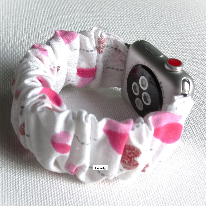 May include: White Apple Watch band with a pink and gray heart pattern. The band is made of a soft, stretchy fabric and has a ruffled design. The text "Lovely" is printed on the band.