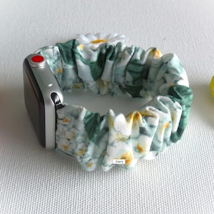 May include: A white and green floral Apple Watch band with a white daisy charm. The band is made of a soft, stretchy fabric and is designed to fit most Apple Watch sizes.