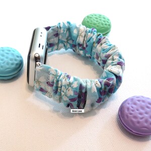 K&ouml;nnte beinhalten: Ein blaues und wei&szlig;es Blumenmuster-Armband aus Stoff f&uuml;r die Apple Watch mit einem silbernen Apple Watch-Zifferblatt. Das Armband besteht aus dehnbarem Stoff und hat einen zerknitterten Look. Der Text "Ann Lee" ist auf dem Bild sichtbar.