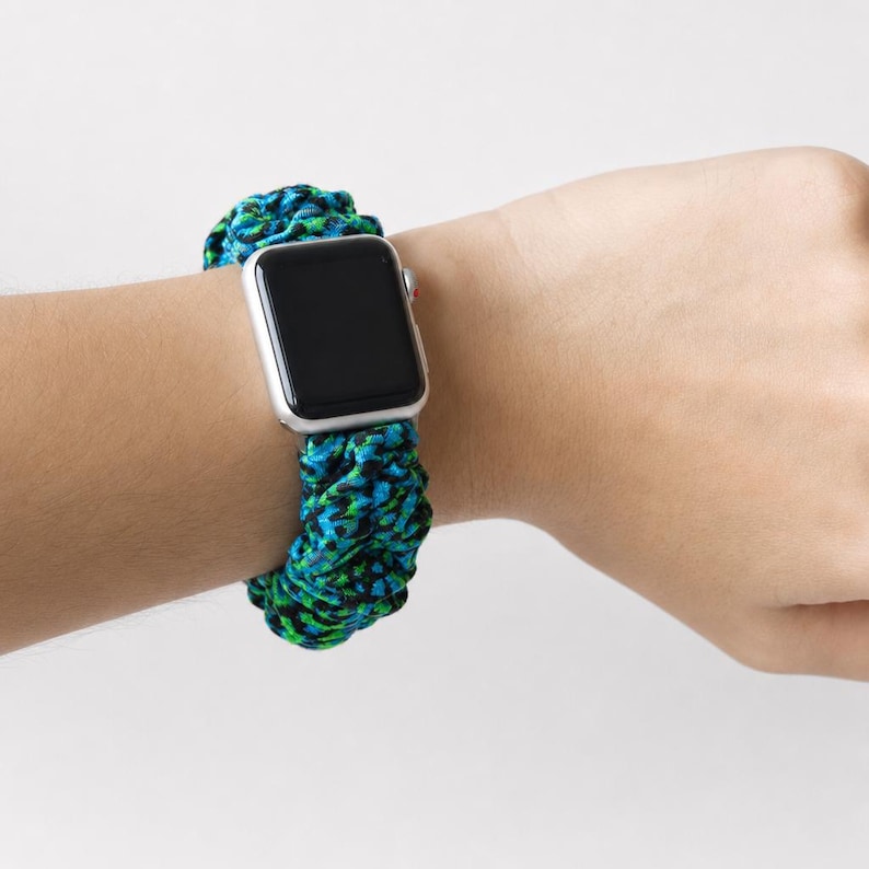 May include: A silver Apple Watch with a black screen is worn on a wrist with a blue, green, and black braided watch band. The band is made of a thick, textured material.
