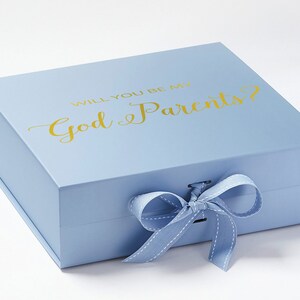 Könnte beinhalten: Eine hellblaue Geschenkbox mit einem weißen Band und dem Text "Willst du mein Pate/meine Patin sein?" in goldenen Buchstaben.