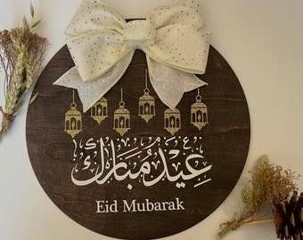 May include: A round wooden sign with a white bow and gold glitter accents. The sign says "Eid Mubarak" in Arabic calligraphy and features a design of gold lanterns.