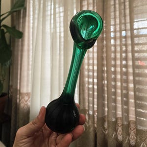 May include: A hand-blown glass vase with a unique, elongated shape. The vase features a deep emerald green color that transitions to a darker, almost black hue at the base. The top has a rounded opening, and the vase is held in a hand.