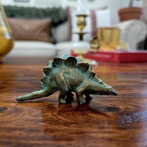 May include: A small, metallic Stegosaurus figurine with a weathered, bronze-toned finish. The dinosaur features a row of prominent, triangular plates along its back, a pointed tail, and four legs positioned on a flat surface. The surface shows patina.