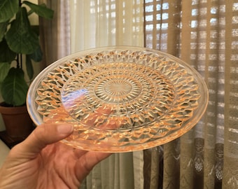 May include: A clear, round glass plate with an intricate, raised geometric pattern in a peach color. The plate has a central circular design and a scalloped edge. The plate is held in a hand.