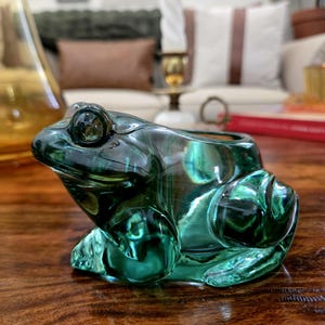 May include: A detailed, emerald green glass frog figurine. The frog is sitting on a wooden surface. The figurine has a large, round eye and a hollowed-out back, possibly for holding small items. The glass has a reflective, glossy finish.