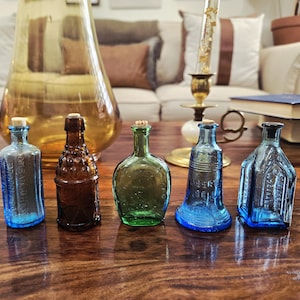 May include: Five antique glass bottles of varying shapes and colors, including blue, brown, and green, with cork stoppers. The bottles are arranged on a dark wooden surface, showcasing their unique designs and textures. A large amber glass vase is in the background.